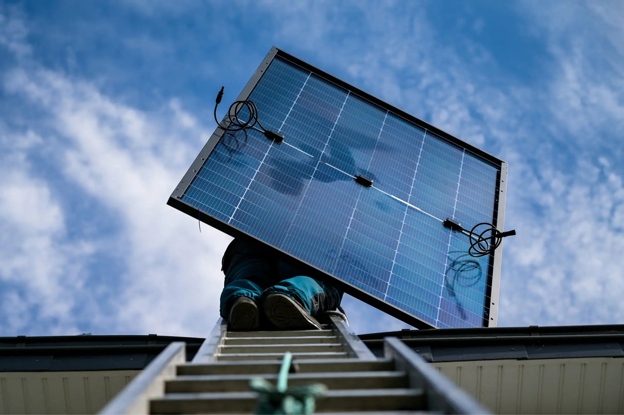 Artisan Sunology HOME portant un panneau solaire photovoltaïque, prêt à être posé sur une toiture en ardoise. L’image montre le professionnel en tenue de travail, avec un ciel clair en arrière-plan, illustrant la pose soignée et experte des modules solaires.